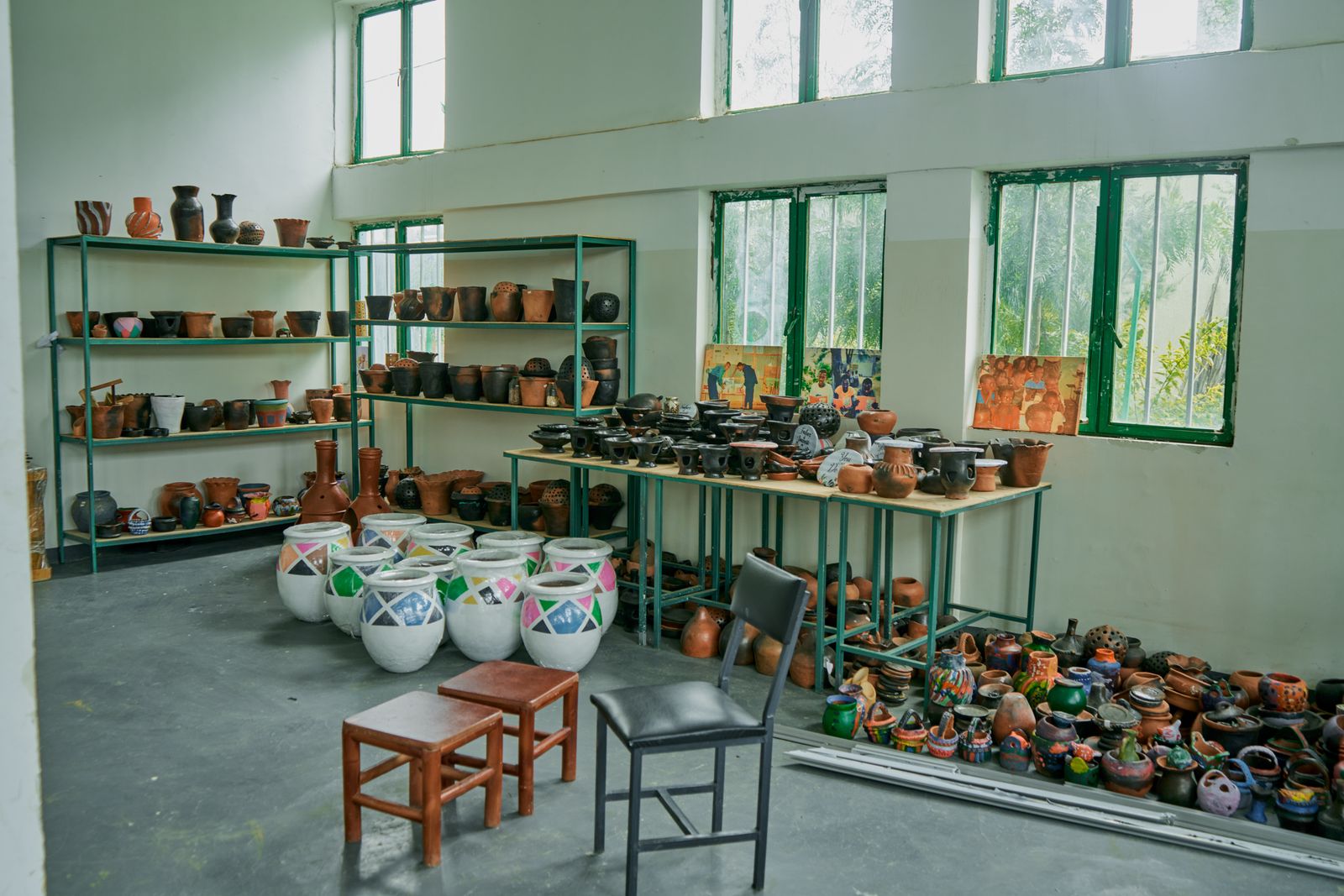 A high-ceilinged ceramics studio with green metal shelving, rows of clay pots on long tables and more pots lined along the floor beside tall windows