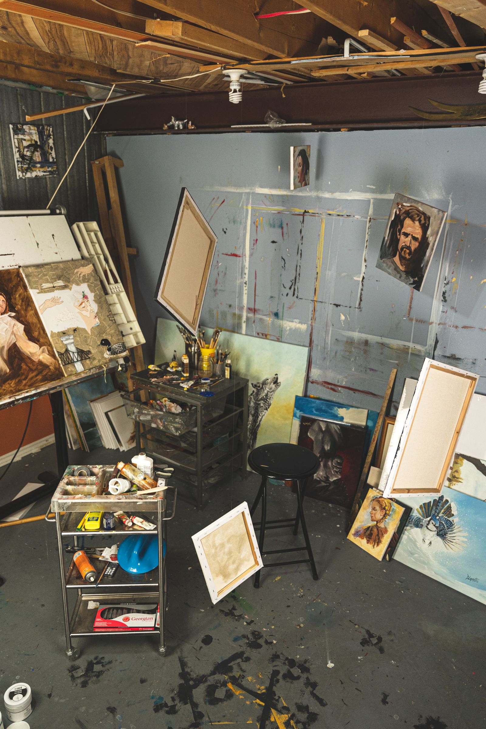 A painter's studio with canvases stacked against a blue wall, an easel, a stool and a rolling cart of paint tubes on a spattered concrete floor