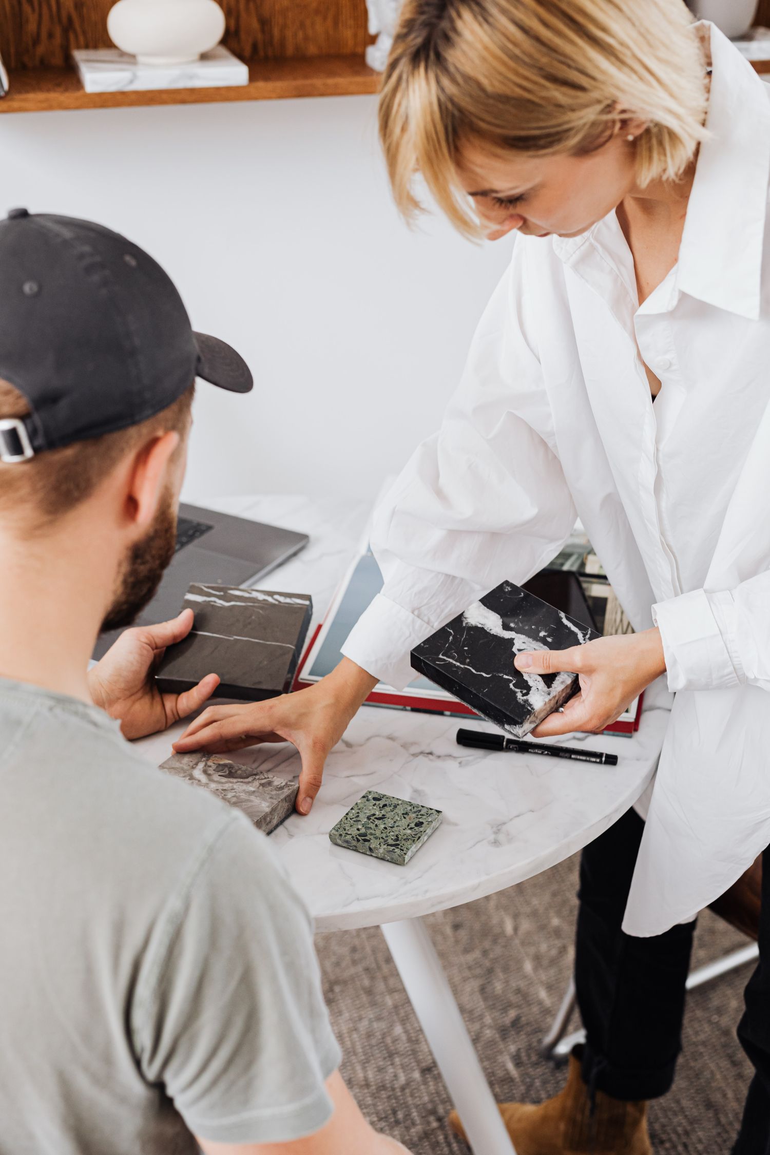 Two people at a round white marble table reviewing stone samples, one holding a black and white marble square while the other rests a hand on a grey sample, with a laptop and tablet nearby