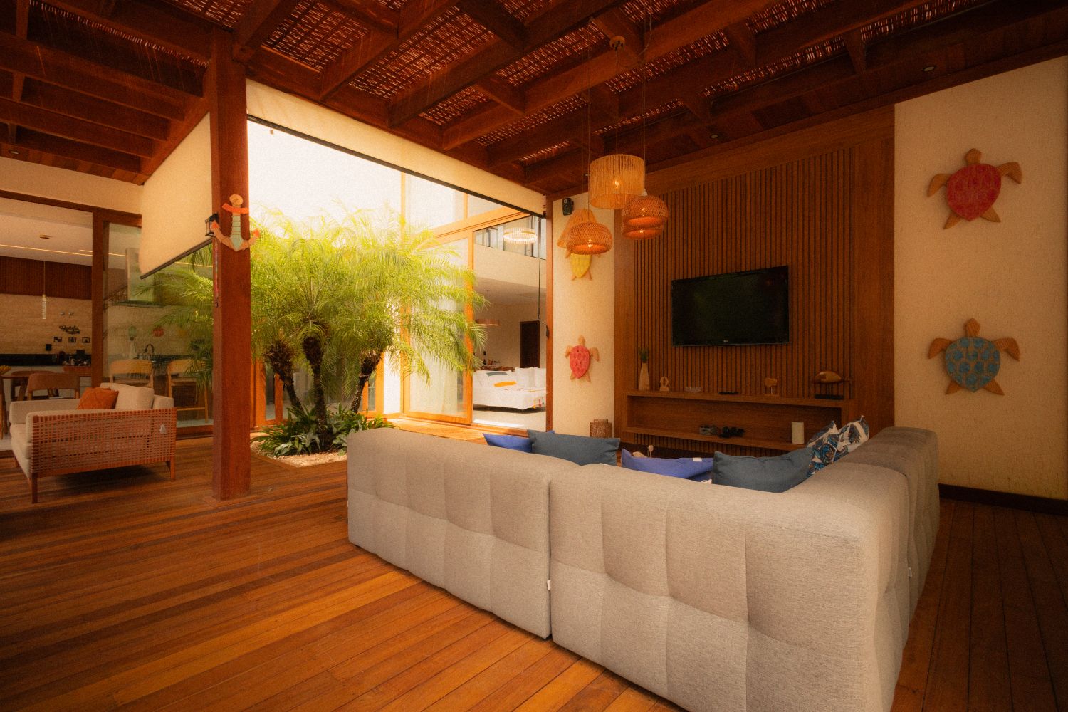 An open-air living room with a warm wood floor and slatted wood ceiling, a long cream sectional sofa with blue cushions, woven pendant lamps and an interior courtyard planted with palms